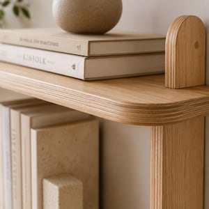 May include: A light-toned wooden wall shelf featuring a smooth, rounded edge design and visible layered plywood construction. The shelf is mounted with a vertical support piece and holds two stacked books and a round, textured decorative object.