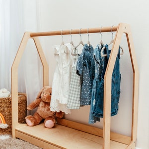 May include: A light-colored wooden clothing rack shaped like a house frame with a horizontal rod. Several small garments hang on hangers from the rod, including a white dress, a checkered dress, and denim items. A brown plush toy sits on the base.