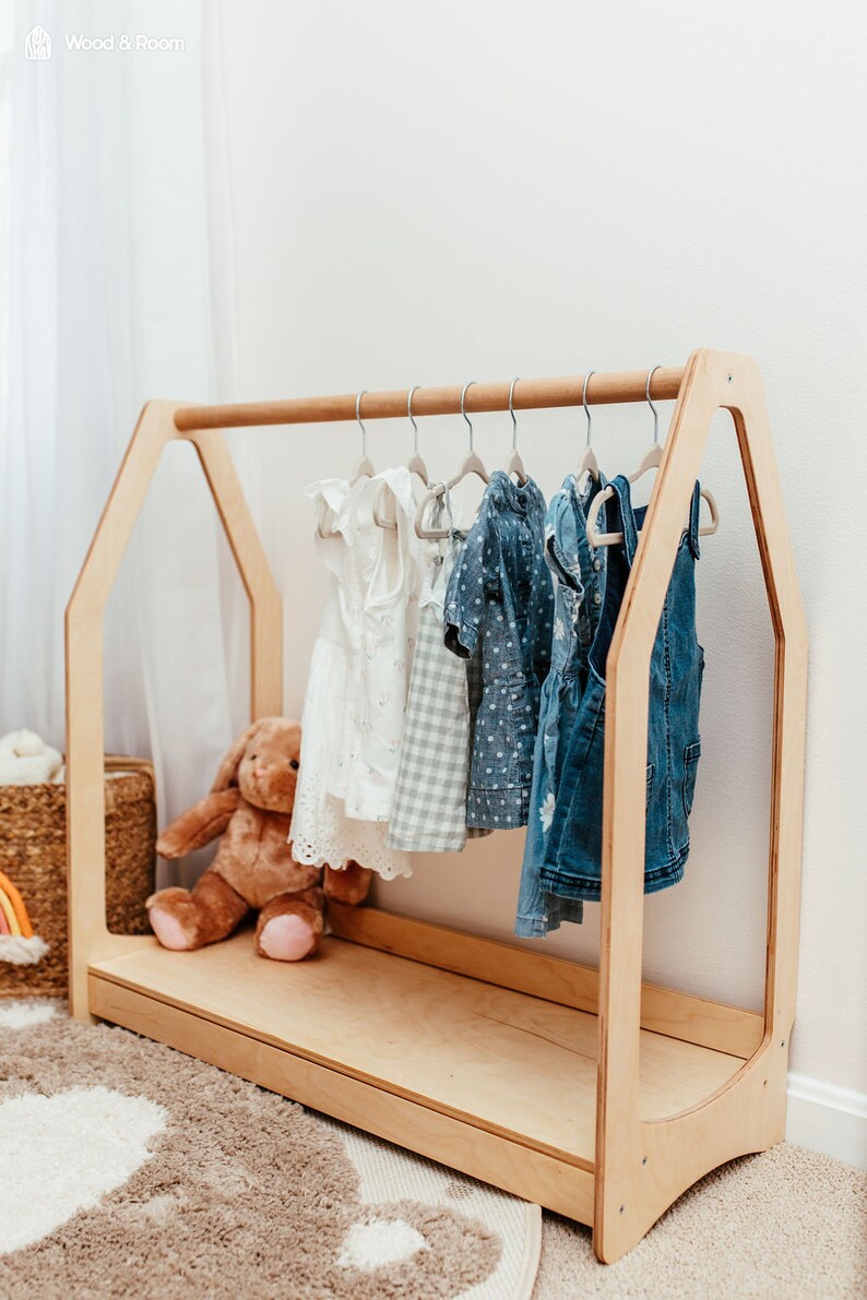 May include: A light-colored wooden clothing rack shaped like a house frame with a horizontal rod. Several small garments hang on hangers from the rod, including a white dress, a checkered dress, and denim items. A brown plush toy sits on the base.