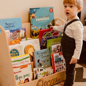 May include: A wooden bookshelf with multiple shelves filled with colorful children's books. The bookshelf is labeled "Oscar" and has a child standing in front of it.
