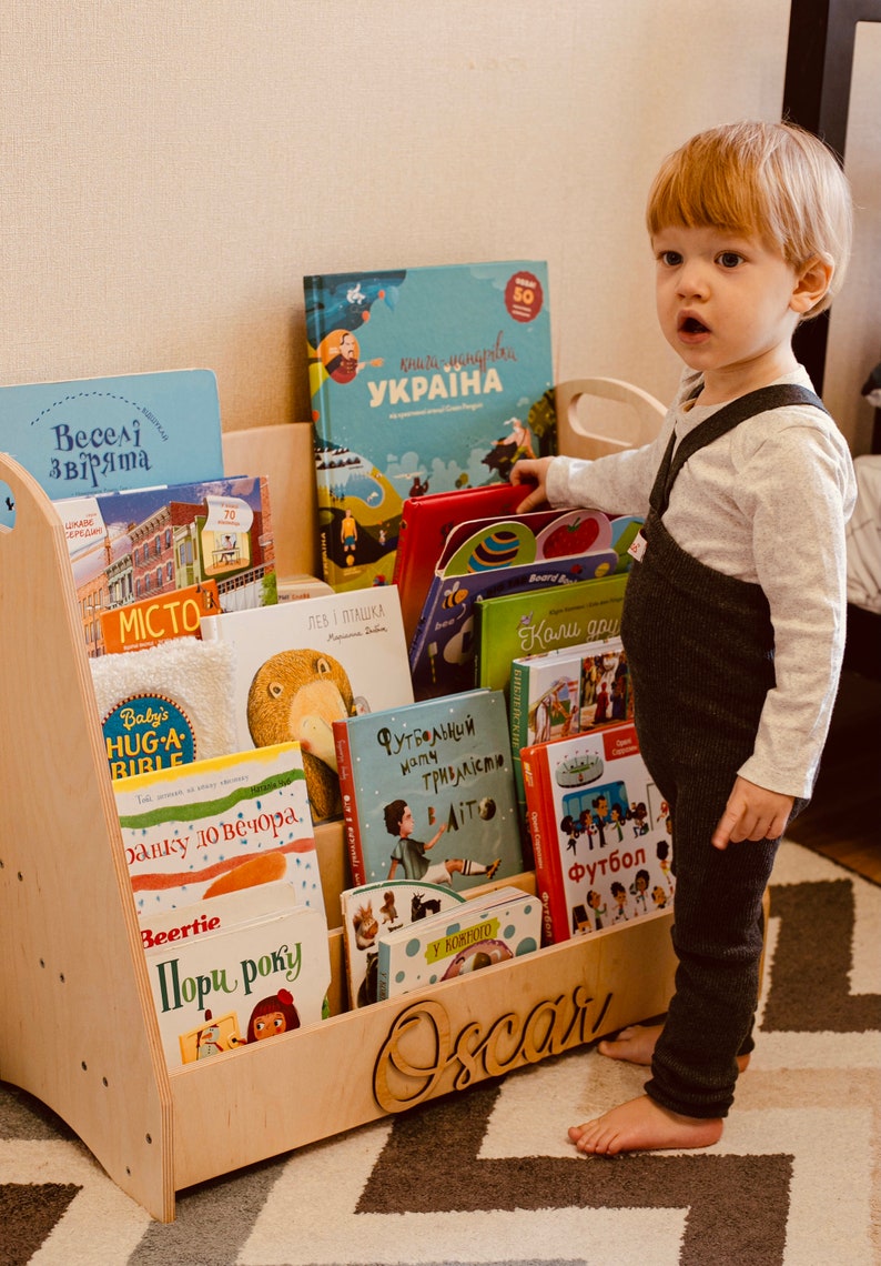 May include: A wooden bookshelf with multiple shelves filled with colorful children's books. The bookshelf is labeled "Oscar" and has a child standing in front of it.