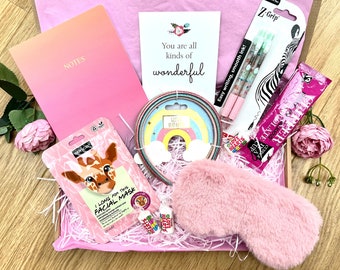 Pode incluir: Uma caixa de presente rosa cheia de itens de autocuidado. Inclui um caderno rosa, uma máscara facial, canetas, uma máscara de dormir, uma faixa de cabelo e um cartão que diz "You are all kinds of wonderful."