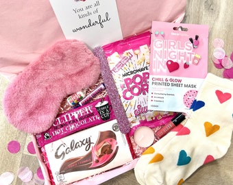 May include: A pink gift box filled with various items, including a pink eye mask, a pink and white Galaxy chocolate bar, a pink and white microwave popcorn bag, a pink face mask with the text "Girls Night In", a pink and white lip gloss, a pink and white candle, and a pair of white socks with colorful hearts.