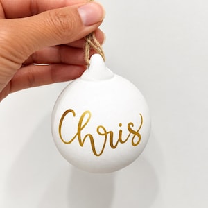 May include: A round white ceramic ornament featuring the name Chris written in gold cursive script. The ornament includes a small loop at the top with a natural brown jute twine string attached for hanging purposes.