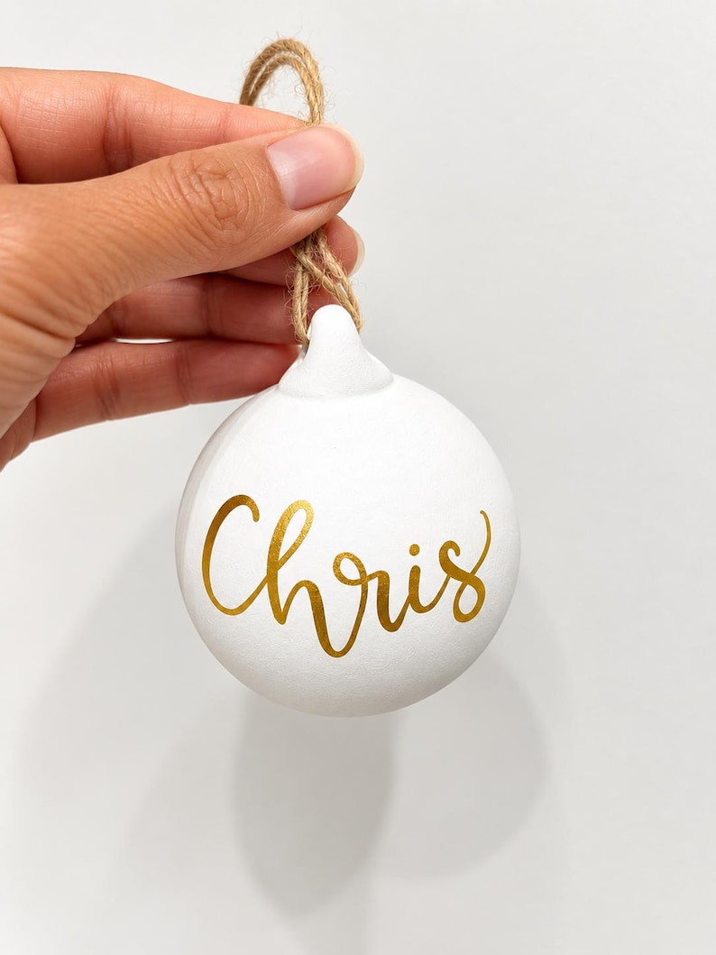 May include: A round white ceramic ornament featuring the name Chris written in gold cursive script. The ornament includes a small loop at the top with a natural brown jute twine string attached for hanging purposes.