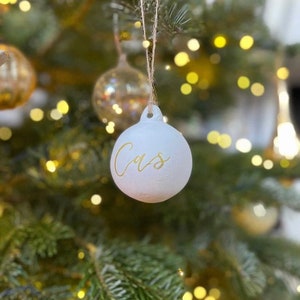 May include: A round, matte white Christmas ornament featuring the name Cas written in elegant gold cursive script. The ornament is suspended by a thin, natural-colored twine string, creating a simple and clean look for holiday tree decoration.