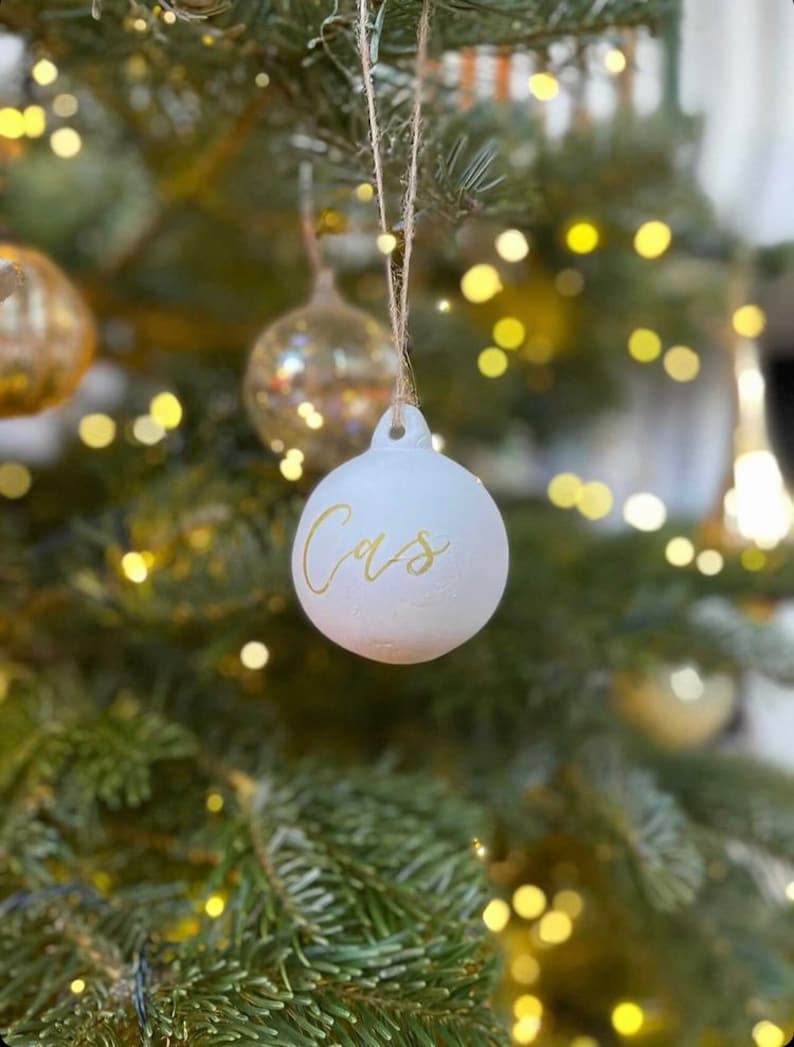 May include: A round, matte white Christmas ornament featuring the name Cas written in elegant gold cursive script. The ornament is suspended by a thin, natural-colored twine string, creating a simple and clean look for holiday tree decoration.