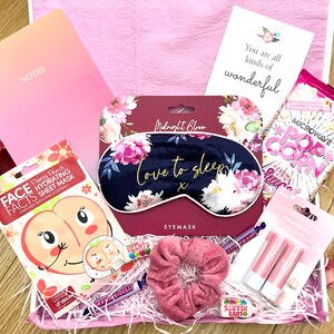 Könnte beinhalten: Eine rosa und weiße Geschenkbox gefüllt mit Selbstpflegeartikeln, darunter eine Blumen-Augenmaske, eine Gesichtsmaske, Lippenbalsam, ein Haargummi und eine Tüte Mikrowellen-Popcorn. Die Box enthält außerdem einen rosa Notizblock mit dem Wort "NOTES" darauf und eine Karte mit der Aufschrift "Du bist einfach wunderbar".