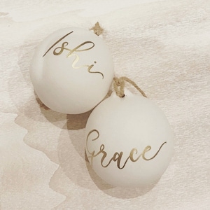 May include: Two round, matte white ornaments featuring gold calligraphy. One ornament displays the name Ishi, and the other displays the name Grace. Each ornament includes a small loop at the top with a piece of natural twine attached for hanging.