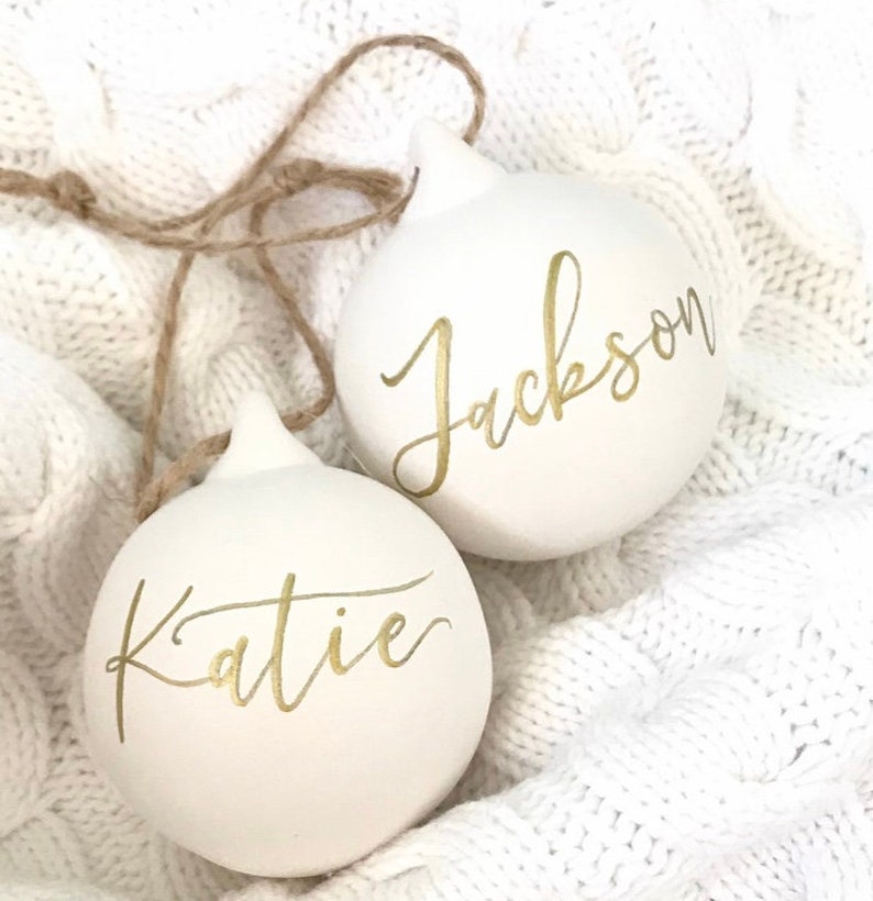 May include: Two round white ornaments with pointed tops and attached brown twine strings. Each ornament features a name written in gold cursive script. One ornament displays the name Katie, and the other ornament displays the name Jackson.