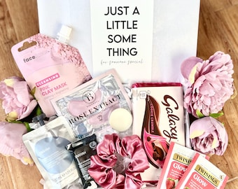May include: A gift box filled with various items, including a pink clay face mask, a rose extract face mask, a white candle, a pink hair scrunchie, a bar of Galaxy chocolate, and two tea bags. The box is decorated with pink flowers and a card that reads "Just a little something for someone special."