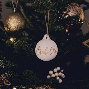 May include: A round white Christmas ornament featuring the name Arabella written in gold cursive script. The ornament has a small loop at the top with a thin brown twine string attached for hanging on a tree branch.