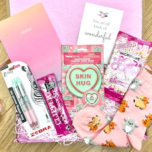 Könnte beinhalten: Eine rosa Geschenkbox gefüllt mit verschiedenen Artikeln, darunter ein rosa Notizbuch mit dem Wort "NOTES" auf dem Cover, zwei rosa Stifte mit Glitzer, eine rosa Gesichtsmaske, eine rosa Tüte Mikrowellenpopcorn und ein Paar rosa Socken mit Katzenmuster. Die Box ist mit rosa und weißem zerknülltem Papier gefüllt. Eine Karte mit dem Text "You are all kinds of wonderful" ist ebenfalls enthalten.