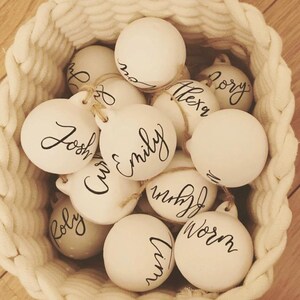 May include: A collection of round, white ceramic ornaments, each featuring a unique name written in black cursive script. The ornaments are suspended by thin twine loops and are contained within a woven, off-white rope basket.
