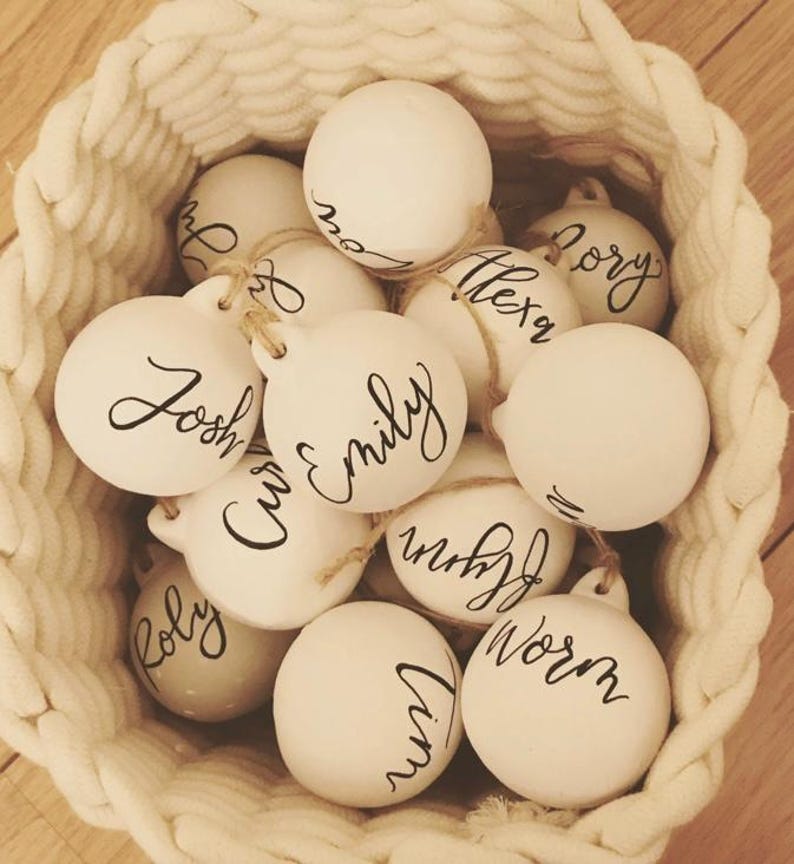 May include: A collection of round, white ceramic ornaments, each featuring a unique name written in black cursive script. The ornaments are suspended by thin twine loops and are contained within a woven, off-white rope basket.