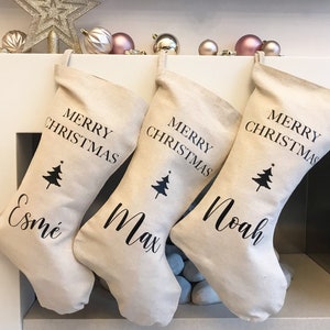 May include: Three beige Christmas stockings with black text reading "MERRY CHRISTMAS" and a Christmas tree graphic. Each stocking is personalised with a name: "Esmé", "Max", and "Noah". Baubles and a star decoration are on the mantelpiece.