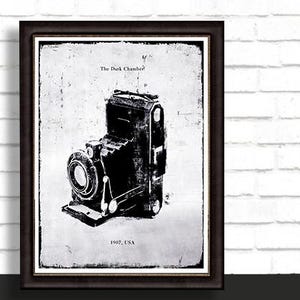 May include: Framed black and white print of a vintage camera. The print features the text "The Dark Chamber" and "1907, USA". The camera is a detailed illustration with a textured background, set in a dark brown frame.