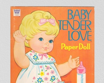 May include: A vintage paper doll of Baby Tender Love, a doll with blonde hair, wearing a pink and white floral dress and white shoes, holding a pink bottle. The paper doll is from a Whitman book.