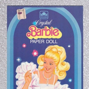 Puede incluir: Un juego de muñecas de papel con Crystal Barbie. La caja muestra una ilustración colorida de Barbie con un vestido blanco y una faja rosa. El juego incluye un soporte de plástico y 14 atuendos precortados. No se necesitan tijeras.