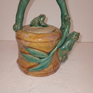 Puede incluir: Una tetera de cerámica con un asa de bambú verde y una rana en la tapa. La tetera está decorada con hojas verdes y tiene un esmalte marrón.