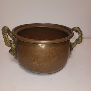 May include: A small copper pot with two ornate brass handles. The pot is round and has a slightly flared rim.