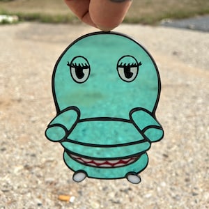 May include: A teal blue cartoon character shaped like an armchair with a toothy grin, large eyes, and eyelashes. The character is made of glass and has a shiny, reflective surface.