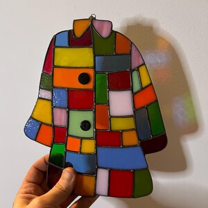 K&ouml;nnte beinhalten: Ein buntes Glasfenster in Form eines Mantels. Es besteht aus rechteckigen und quadratischen Glasst&uuml;cken in Rot, Gelb, Orange, Blau, Gr&uuml;n und Rosa. Zwei schwarze runde Kn&ouml;pfe sind sichtbar. Dekoration.