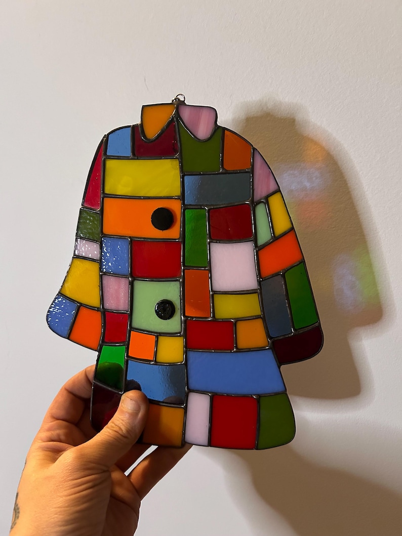 K&ouml;nnte beinhalten: Ein buntes Glasfenster in Form eines Mantels. Es besteht aus rechteckigen und quadratischen Glasst&uuml;cken in Rot, Gelb, Orange, Blau, Gr&uuml;n und Rosa. Zwei schwarze runde Kn&ouml;pfe sind sichtbar. Dekoration.