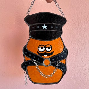 May include: Stained glass art piece featuring an orange figure with a black hat, mustache, and harness. The hat has a white star. A silver chain is attached for hanging. The figure has a whimsical, cartoonish design.