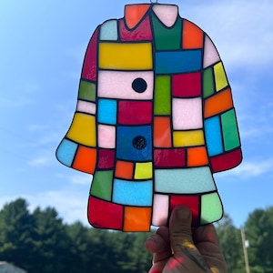 K&ouml;nnte beinhalten: Ein bunter, mantelformiger Glas-Suncatcher. Das Design zeigt ein Patchwork aus leuchtenden Farben wie Rot, Gelb, Blau, Gr&uuml;n und Rosa mit schwarzen Akzenten. Der Suncatcher wird vor einem blauen Himmel gehalten.