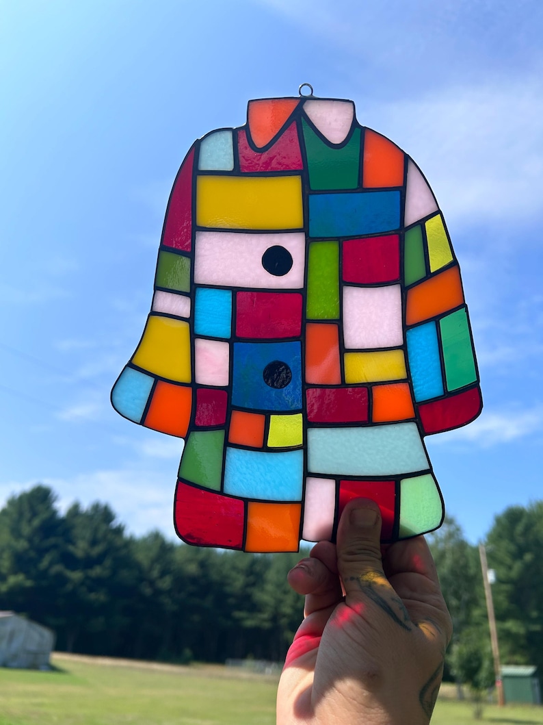 K&ouml;nnte beinhalten: Ein bunter, mantelformiger Glas-Suncatcher. Das Design zeigt ein Patchwork aus leuchtenden Farben wie Rot, Gelb, Blau, Gr&uuml;n und Rosa mit schwarzen Akzenten. Der Suncatcher wird vor einem blauen Himmel gehalten.