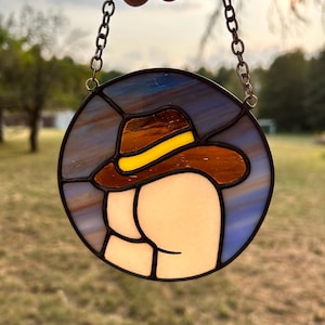 May include: A stained glass suncatcher featuring a cartoon-style rear end wearing a brown cowboy hat with a yellow band. The suncatcher is round and has a blue and brown background.