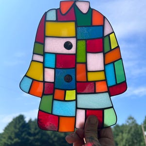 K&ouml;nnte beinhalten: Ein buntes Glasbild in Form eines Mantels mit einem Patchwork-Design aus verschiedenen Farben wie Rot, Gelb, Blau und Gr&uuml;n. Das St&uuml;ck hat einen schwarzen Rand und wird vor einem blauen Himmel gehalten.