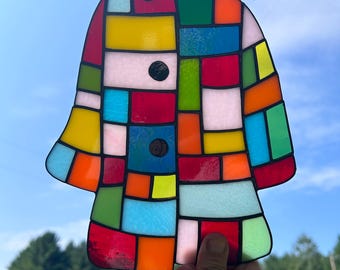 May include: A colourful stained glass coat-shaped artwork. The design is a patchwork of red, yellow, blue, and green glass pieces, outlined in black. The artwork is held up against a blue sky, with trees visible in the background.
