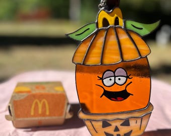 May include: A stained glass pumpkin ornament with a smiling face and a jack-o'-lantern face at the bottom. The pumpkin is orange with green leaves and a yellow stem.