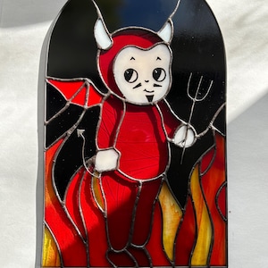 May include: A stained glass window depicting a cartoon devil in a red suit with a white face and large eyes. The devil is standing in flames and holding a pitchfork.