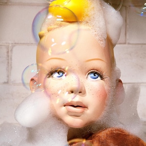 May include: A child's doll head with a yellow rubber ducky on top, covered in white foam bubbles. The doll has blue eyes, freckles, and pink cheeks. There are several soap bubbles in the background.