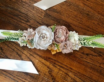 May include: A white satin sash with a floral embellishment. The embellishment features a large white flower with pearl accents, smaller blush pink flowers, and gold accents.