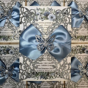 Puede incluir: Una invitaci&oacute;n de Quincea&ntilde;era azul y plateada con un lazo de sat&eacute;n azul y mariposas de brillantina plateada. La invitaci&oacute;n incluye el texto "por haberme permitido llegar a esta vida. Llena de ilusi&oacute;n en mi pady" y "Me los 15". La invitaci&oacute;n tambi&eacute;n incluye la fecha, la hora y la ubicaci&oacute;n del evento: "Comida: 5:00-7:00PM", "Baile: 7:00PM-12:00AM", "St. Paul Armenian Church", "7 N First St. Fresno CA, 93701".