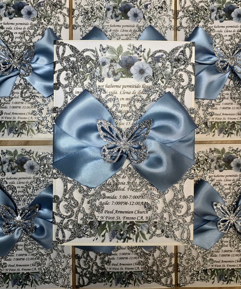 Puede incluir: Una invitaci&oacute;n de Quincea&ntilde;era azul y plateada con un lazo de sat&eacute;n azul y mariposas de brillantina plateada. La invitaci&oacute;n incluye el texto "por haberme permitido llegar a esta vida. Llena de ilusi&oacute;n en mi pady" y "Me los 15". La invitaci&oacute;n tambi&eacute;n incluye la fecha, la hora y la ubicaci&oacute;n del evento: "Comida: 5:00-7:00PM", "Baile: 7:00PM-12:00AM", "St. Paul Armenian Church", "7 N First St. Fresno CA, 93701".