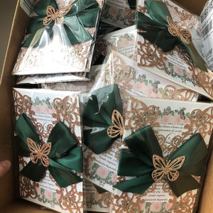 Puede incluir: Una pila de invitaciones de brillo dorado y cinta verde con un dise&ntilde;o de mariposa. Las invitaciones tienen el texto "Sonido Inquieto" e "Invitaci&oacute;n Requerida".