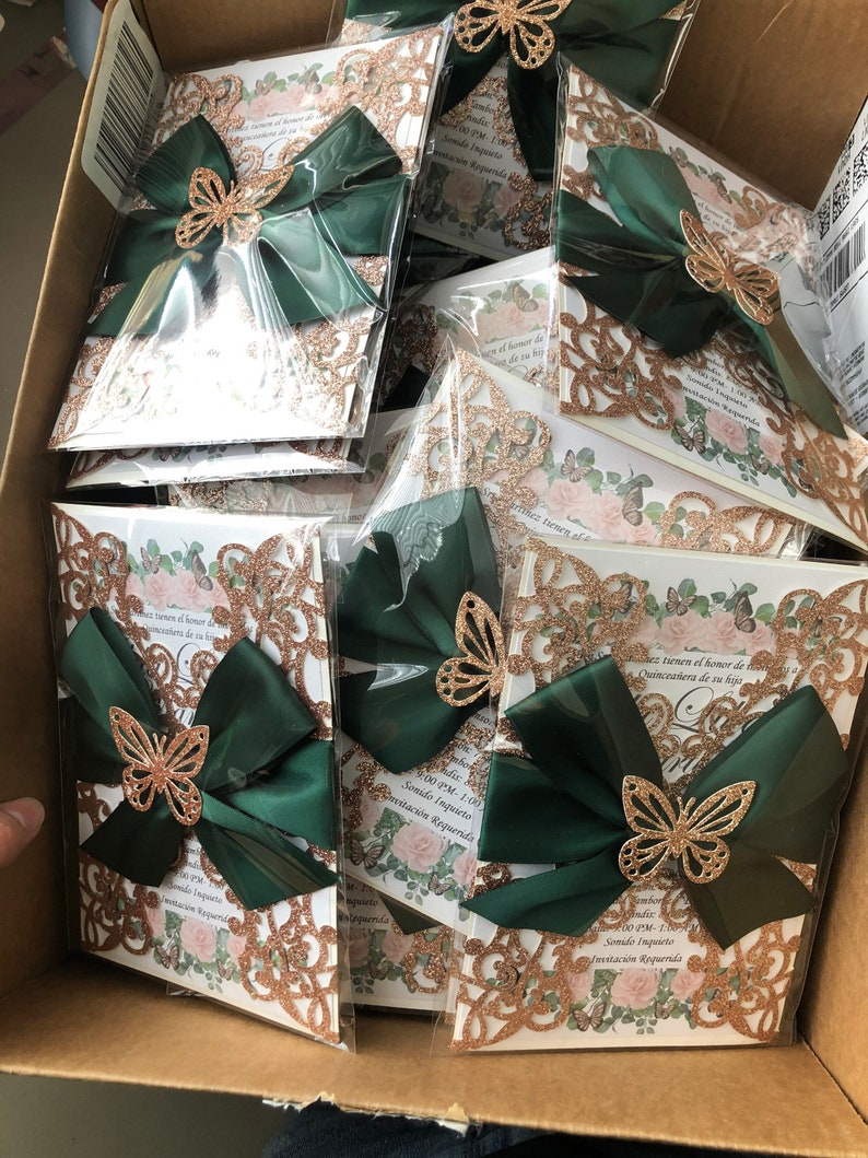 Puede incluir: Una pila de invitaciones de brillo dorado y cinta verde con un dise&ntilde;o de mariposa. Las invitaciones tienen el texto "Sonido Inquieto" e "Invitaci&oacute;n Requerida".