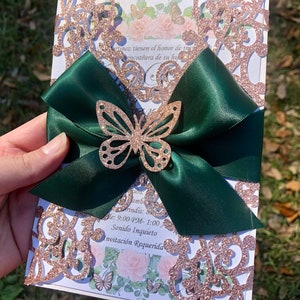 Puede incluir: Una invitaci&oacute;n de corte l&aacute;ser con brillo de oro rosa, un lazo de sat&eacute;n verde y un acento de mariposa de brillo dorado. El texto de la invitaci&oacute;n dice "Martinez tienen el honor de invitar a la quincea&ntilde;era de su hija", "Amb", "Brindis", "Hora: 9:00 PM - 1:00", "Sonido Inquieto", "Invitaci&oacute;n Requerida".