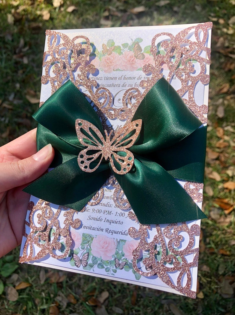Puede incluir: Una invitaci&oacute;n de corte l&aacute;ser con brillo de oro rosa, un lazo de sat&eacute;n verde y un acento de mariposa de brillo dorado. El texto de la invitaci&oacute;n dice "Martinez tienen el honor de invitar a la quincea&ntilde;era de su hija", "Amb", "Brindis", "Hora: 9:00 PM - 1:00", "Sonido Inquieto", "Invitaci&oacute;n Requerida".