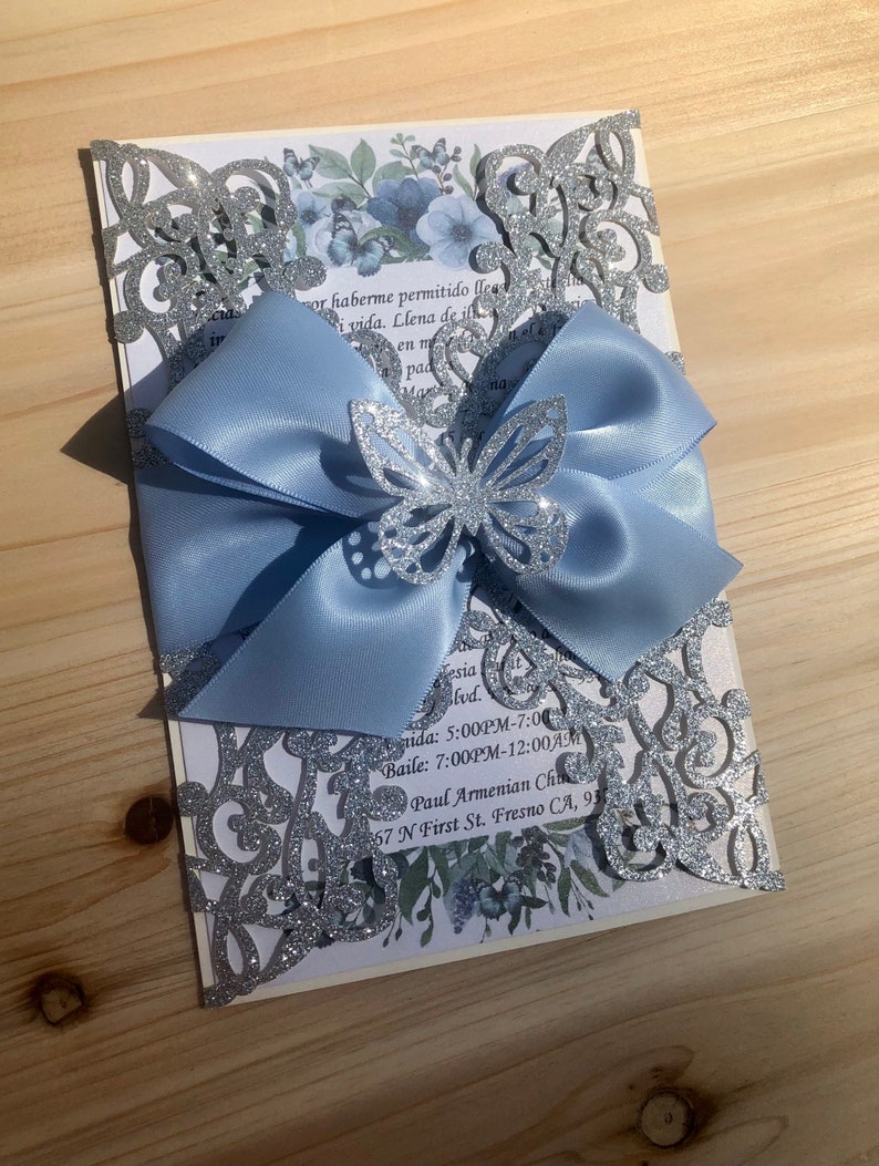 Puede incluir: Una invitaci&oacute;n de corte l&aacute;ser con brillo plateado, un lazo de sat&eacute;n azul claro y una mariposa de brillo plateado. El texto de la invitaci&oacute;n dice: "mor haberme permitido llea i vida. Llena de in en m pad nd esia lvd. ida: 5:00PM-7:00 Baile: 7:00PM-12:00AM Paul Armenian Cha 57 N First St. Fresno CA, 93"