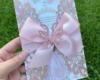 May include: A pink and gold glitter laser cut invitation with a large pink satin bow and a gold glitter butterfly. The invitation has a floral design and the text "Hiro y Amalia Aqui Te Invitan a Celebrar"