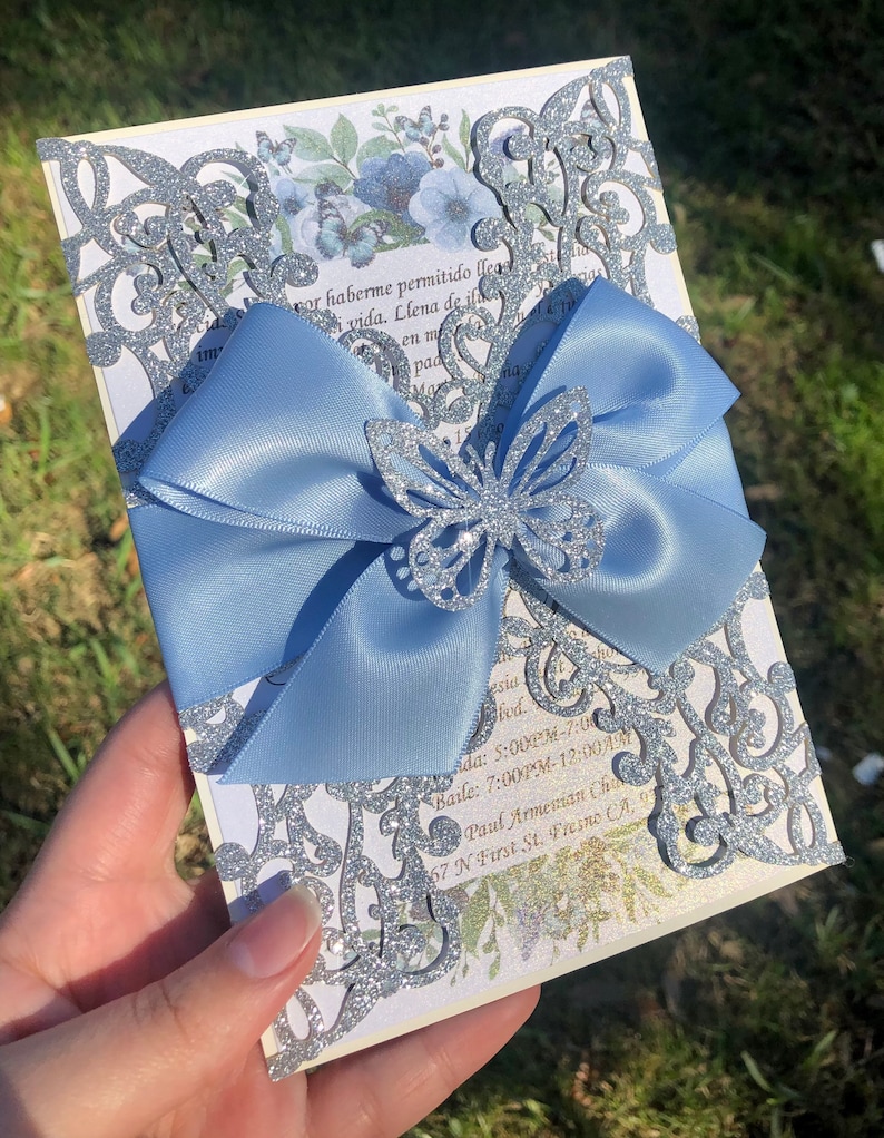 Puede incluir: Una invitaci&oacute;n de corte l&aacute;ser con brillo plateado, un lazo de sat&eacute;n azul claro y una mariposa de brillo plateado. La invitaci&oacute;n tiene un dise&ntilde;o floral y el texto "por haberme permitido llegar a esta vida. Llena de ilusiones en mi  pac sid lvd. Add: 5:00PM-7:00 Baile: 7:00PM-12:00 AM Paul Armessian Church 67 N First St. Fresno CA, "
