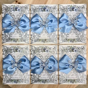 Puede incluir: Seis invitaciones de color plateado con brillo y un patr&oacute;n floral, cada una con un lazo de sat&eacute;n azul claro y una mariposa de brillo plateado. Las invitaciones tienen el texto "por haberme permitido llegar a esta vida. Llena de salud y amor en mi  quincea&ntilde;era" y "Fecha: 17:00 - 19:00  Recepci&oacute;n: 19:00 - 00:00  Iglesia Paul Amensan  27 First St. Fresno, CA 93721"
