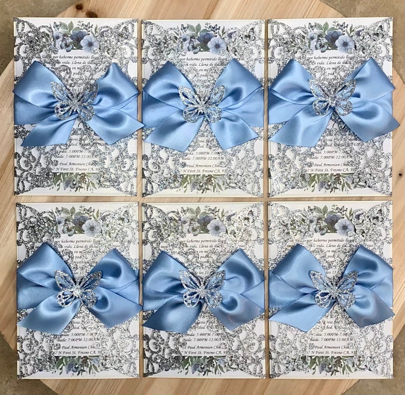 Puede incluir: Seis invitaciones de color plateado con brillo y un patr&oacute;n floral, cada una con un lazo de sat&eacute;n azul claro y una mariposa de brillo plateado. Las invitaciones tienen el texto "por haberme permitido llegar a esta vida. Llena de salud y amor en mi  quincea&ntilde;era" y "Fecha: 17:00 - 19:00  Recepci&oacute;n: 19:00 - 00:00  Iglesia Paul Amensan  27 First St. Fresno, CA 93721"