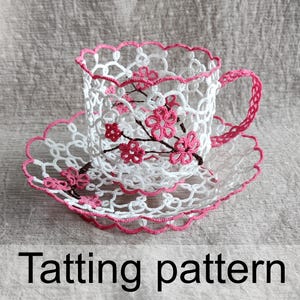 May include: A white and pink tatting pattern teacup and saucer set. The teacup and saucer are intricately crocheted with a floral design. The rim and handle of the teacup are trimmed in pink. The text "Tatting pattern" is at the bottom.